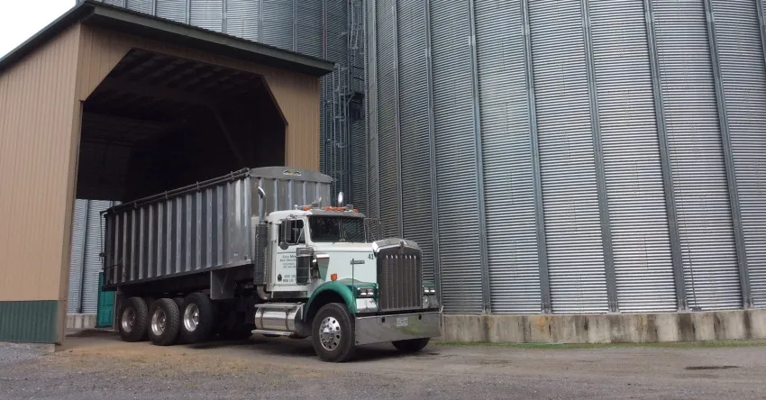 Semi-truck with a large grain trailer parked beside tall metal grain storage bins at an agricultural facility.