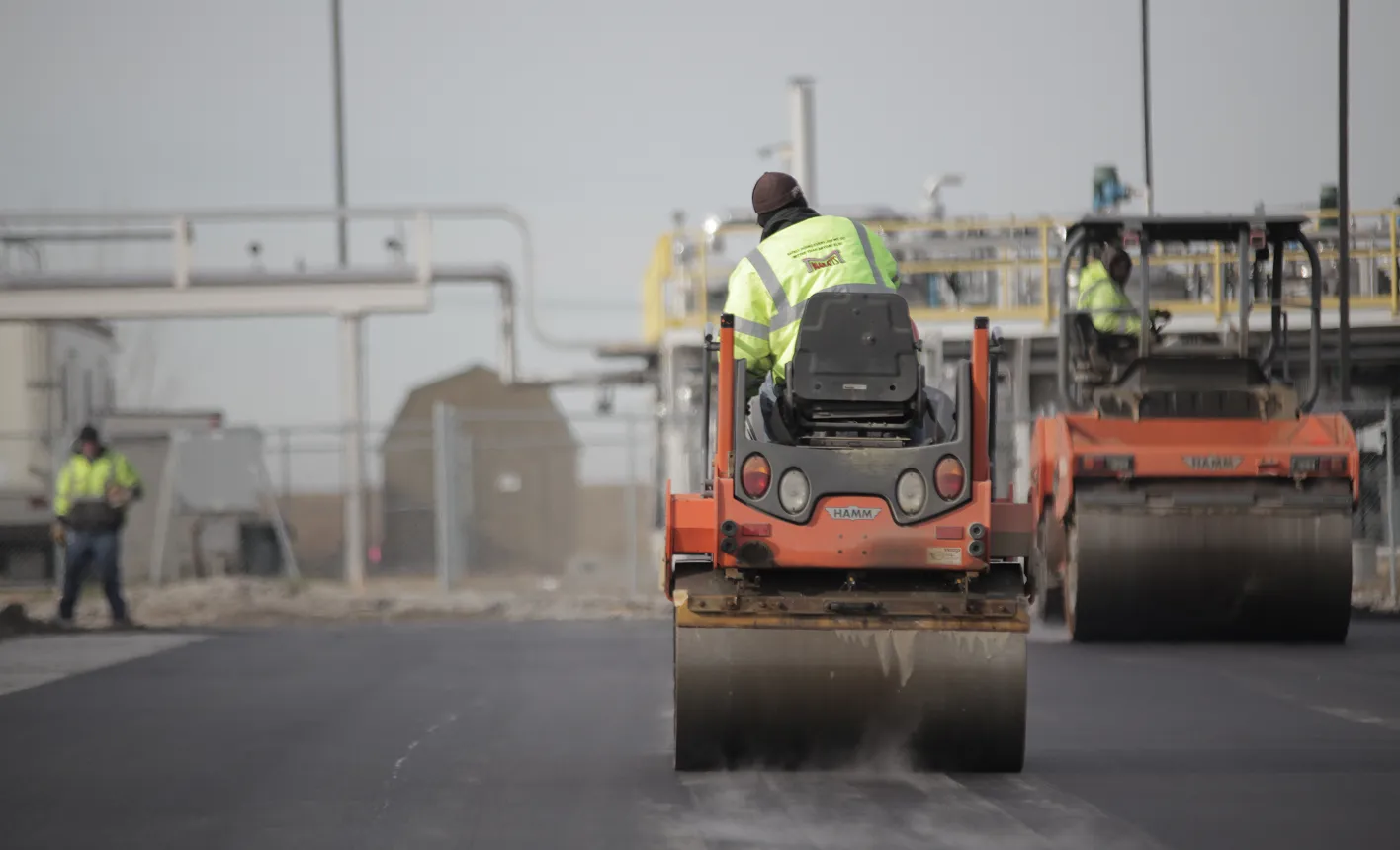 Road rollers compacting freshly paved asphalt on a roadway, with construction workers and equipment visible in the background.