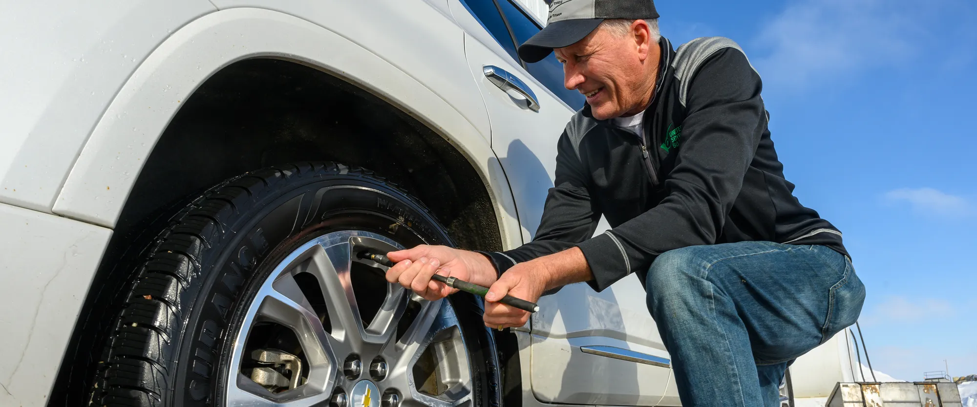 Man kneeling beside a vehicle and checking the tire pressure with a handheld gauge.
