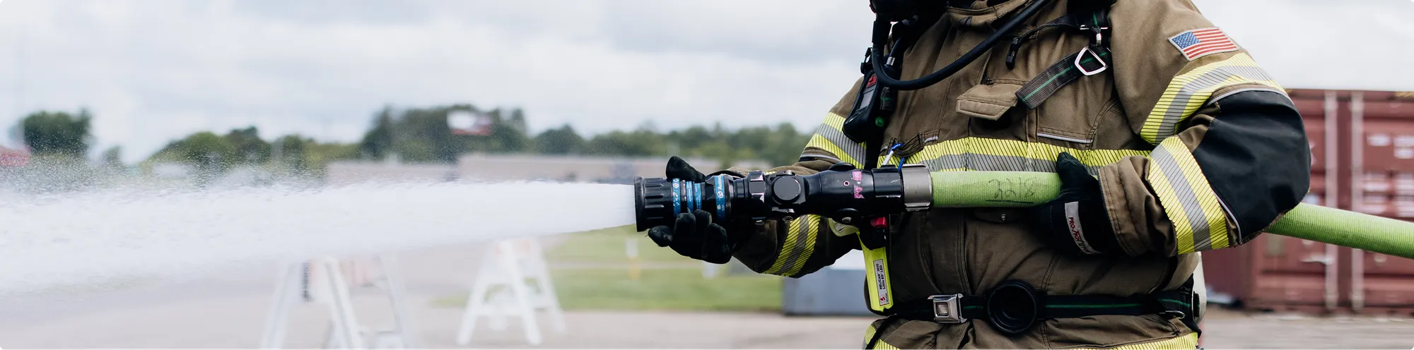 Firefighter in protective gear operating a high-pressure hose, spraying soy-based firefighting foam across an outdoor area.