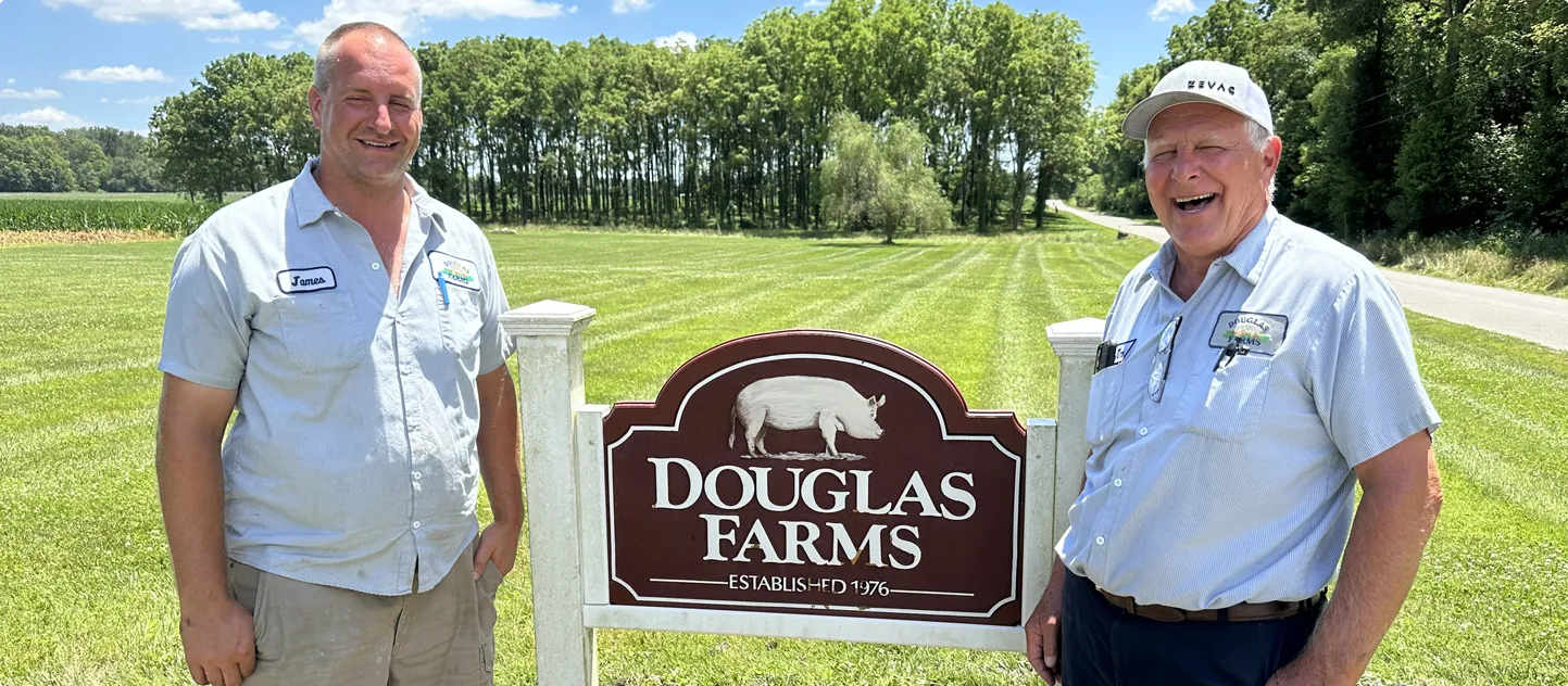 Two Douglas Farms employees laughing beside the farm's wooden entrance sign, with green fields and trees behind them.