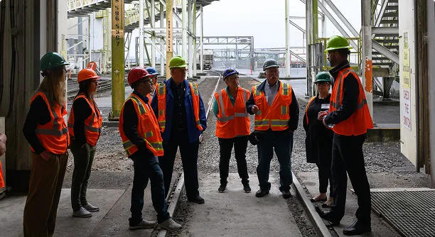 Group of people wearing protective equipment touring an industrial processing facility, with large-scale equipment and infrastructure visible in the background.