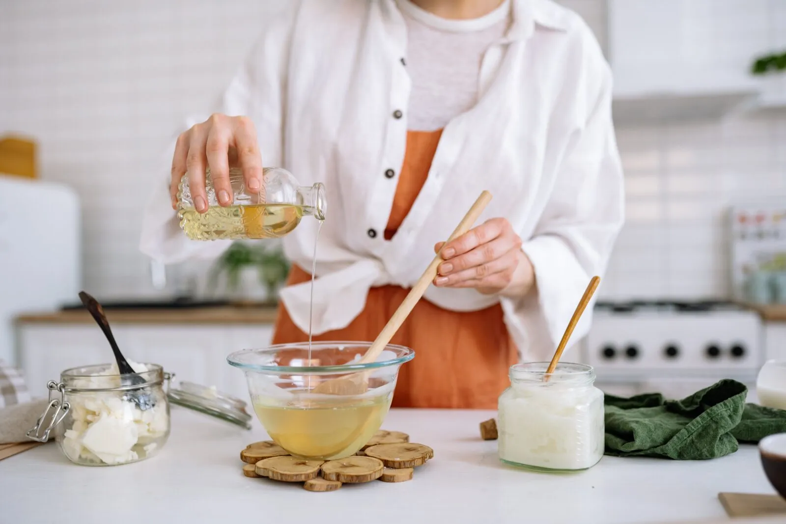 Person pouring oil into a glass bowl while stirring ingredients on a kitchen counter, with jars of homemade products nearby.