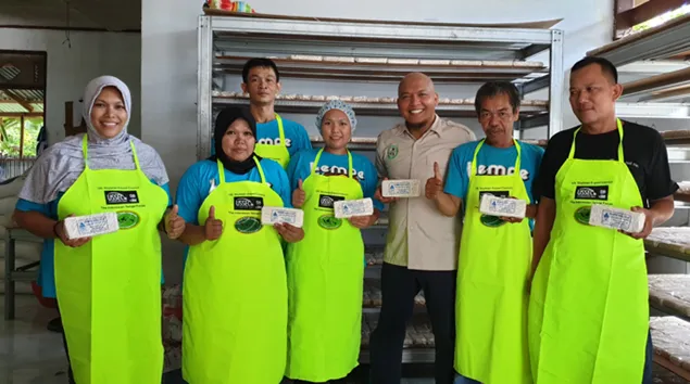 Group of six workers wearing bright green aprons stand inside a food production facility with another man, holding packaged products and giving thumbs-up gestures in front of metal storage racks.