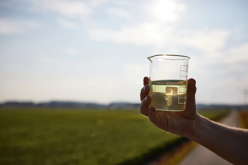 Hand holding a beaker of pale yellow liquid in front of a green agricultural field.