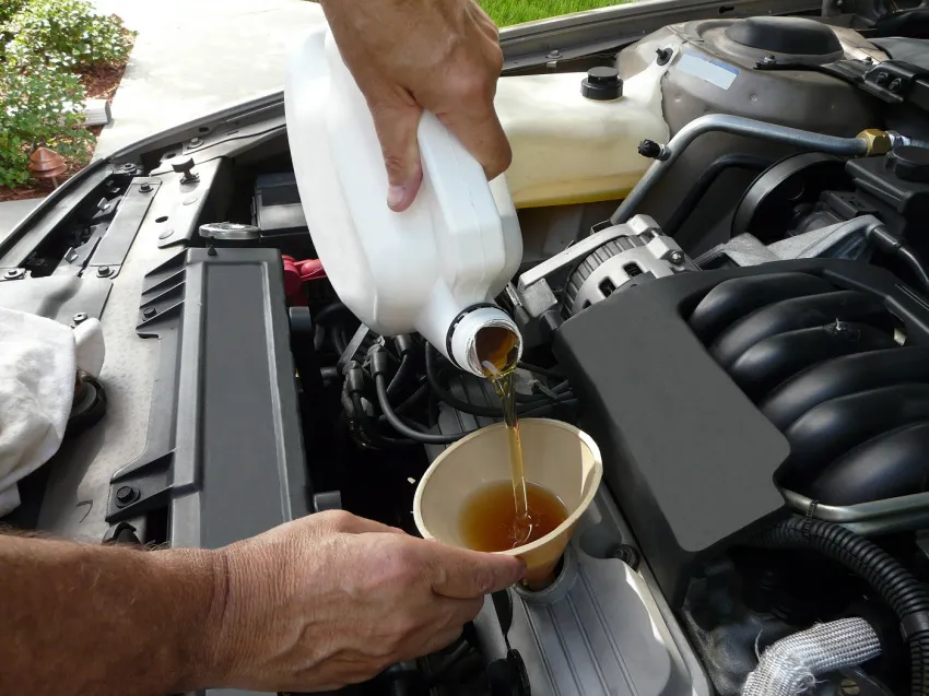 Close-up of a person pouring motor oil from a white container into a funnel positioned over an open car engine.