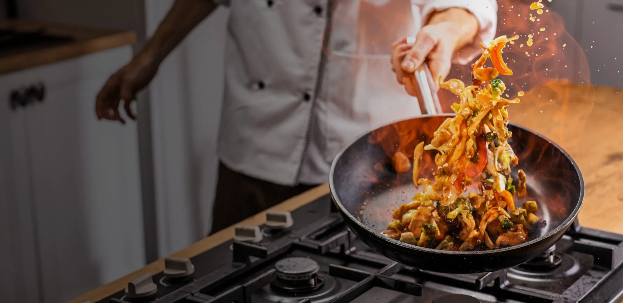 A chef tosses vegetables and noodles in a flaming skillet on a stovetop.