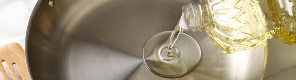 Cooking oil being poured into a stainless steel skillet, with kitchen utensils and a countertop visible.