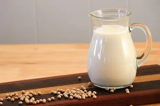 Glass pitcher of soy milk on a wooden table, with scattered soybeans and a wooden serving board in the foreground.