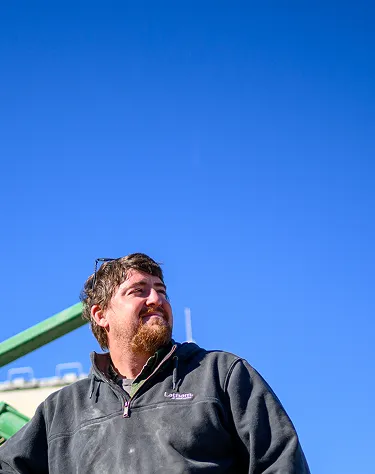 Farmer standing outside near farm equipment with an American flag visible against a blue sky.