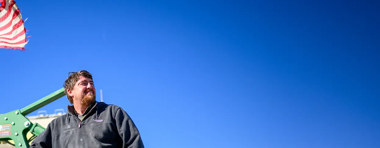 Farmer standing outside near farm equipment with an American flag visible against a blue sky.
