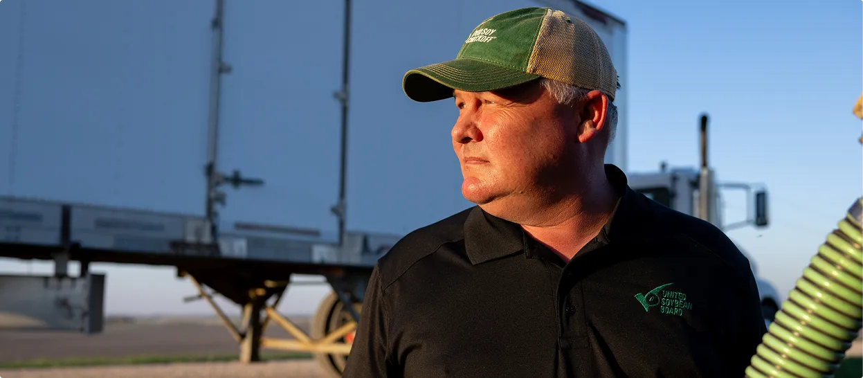 Farmer-leader Tom Frisch standing outdoors near a grain trailer and farm equipment, looking into the distance.