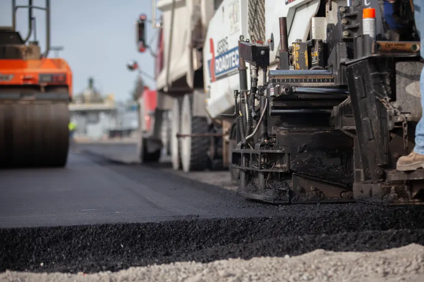 Close-up of asphalt paving equipment laying fresh black pavement on a roadway, with a road roller and additional construction equipment in the background.