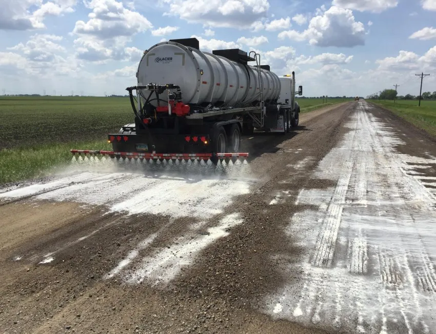 Tanker truck sprays a soy-based dust suppressant across an unpaved rural road bordered by agricultural fields under a partly cloudy sky.