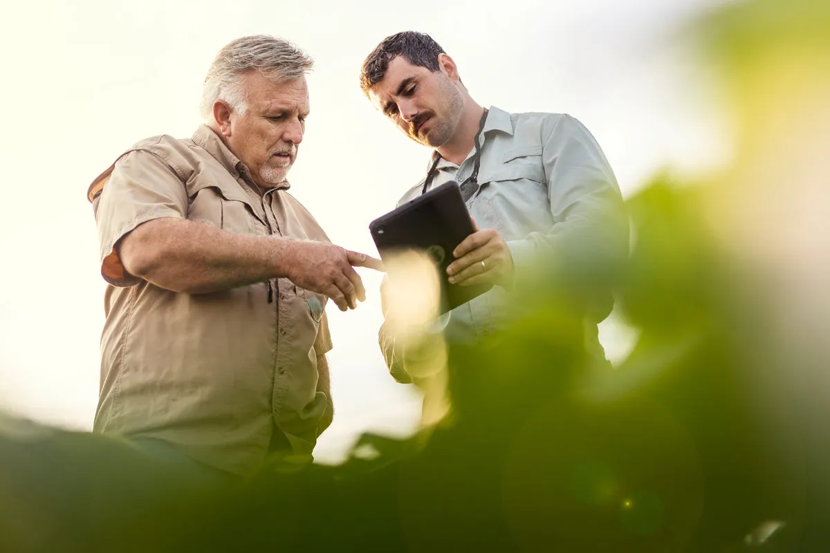 Two men standing in a field review information together on a tablet.
